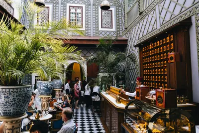 Guests and servers inside an ornate Marrakech coffee house with palm pots and checkered floors