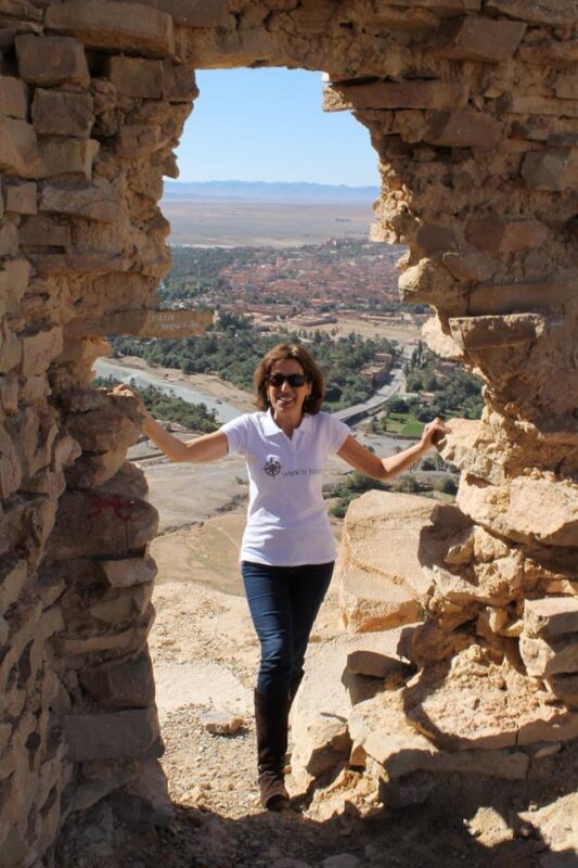 Alexandra Rego smiling in sunglasses, standing in a stone ruin overlooking a palm oasis and desert valley