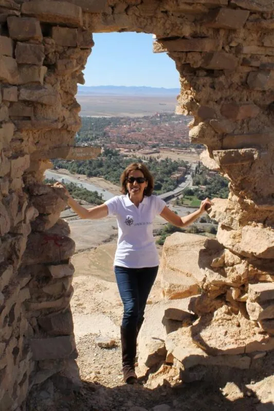 Alexandra Rego smiling in sunglasses, standing in a stone ruin overlooking a palm oasis and desert valley