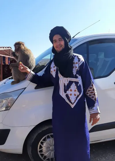 Jamal Ouzina in a blue embroidered robe and black turban offering food to a Barbary macaque on a van hood