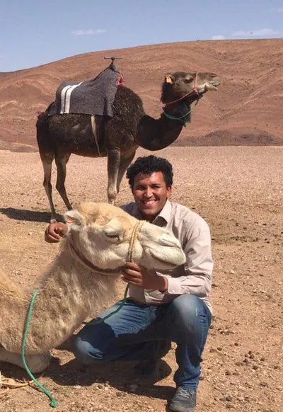 Mohamed Boughrara kneeling in the desert, arms around a cream camel with another saddled camel behind