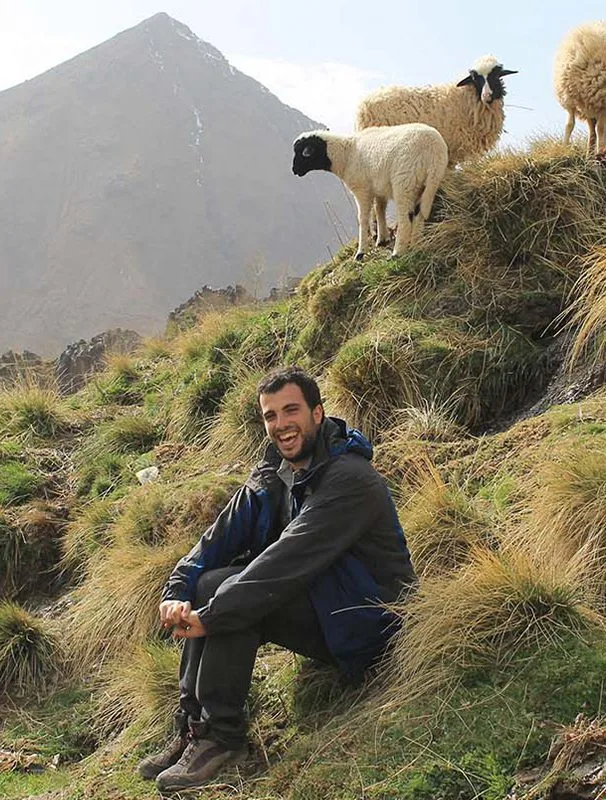 Pedro Lencastre smiling on a grassy mountain slope with black-faced sheep and a pyramid peak behind him