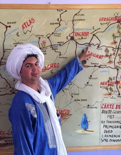 Rachid Ouzina in a blue robe and white turban pointing to Tinjdad on a hand-painted map of southern Morocco