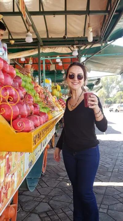 Sónia Medeiros holding a fresh juice beside a yellow market cart stacked with pomegranates and citrus