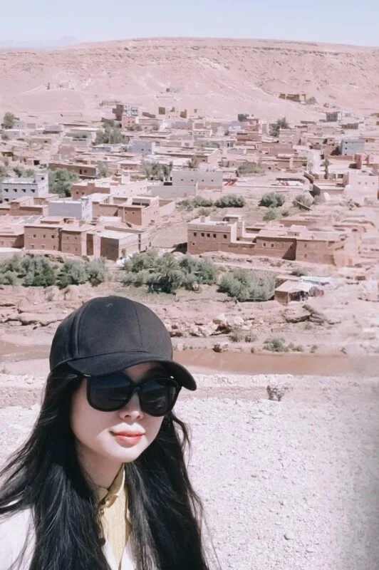 Thea Xu in a black cap and sunglasses selfie above a terracotta desert town nestled under a flat plateau