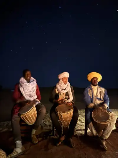 Three men in colourful robes playing drums under a star-filled Sahara night sky