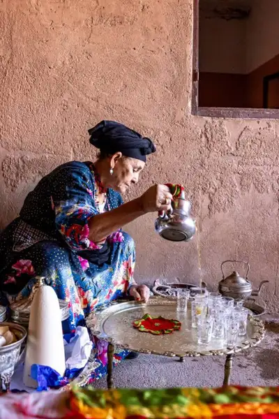 Woman in traditional dress pouring mint tea from a silver pot onto a brass tray