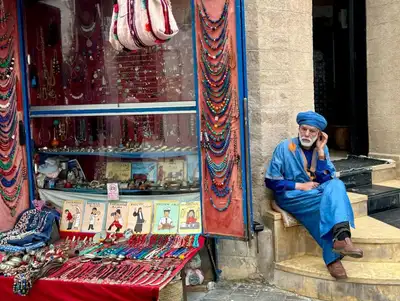 Man in a blue turban and robe sitting beside a shop window packed with beaded jewellery