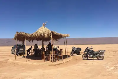 Travellers resting under a thatched shelter beside motorbikes and a 4x4 in the open desert