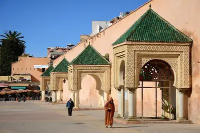 Man in a brown djellaba walking past green-tiled pavilions in a sunlit city square