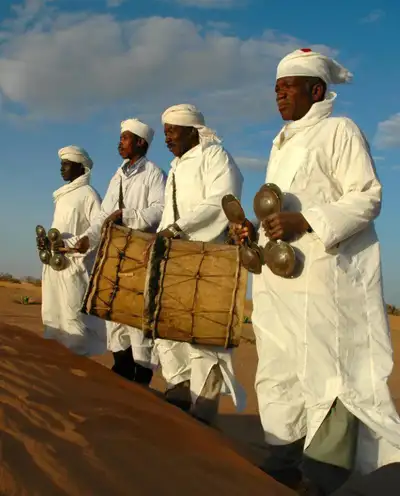 Gnaoua musicians in white robes playing a drum and metal qraqeb castanets on a desert dune