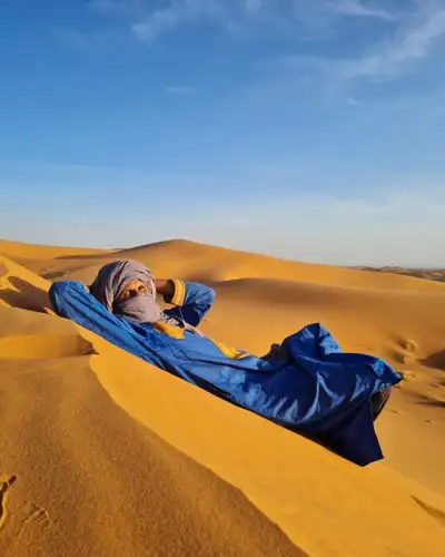 Man in a blue robe and tagelmust reclining on a golden sand dune under a clear sky