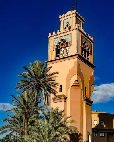 Mosque minaret with carved arches and green tilework rising above palm trees against a clear blue sky