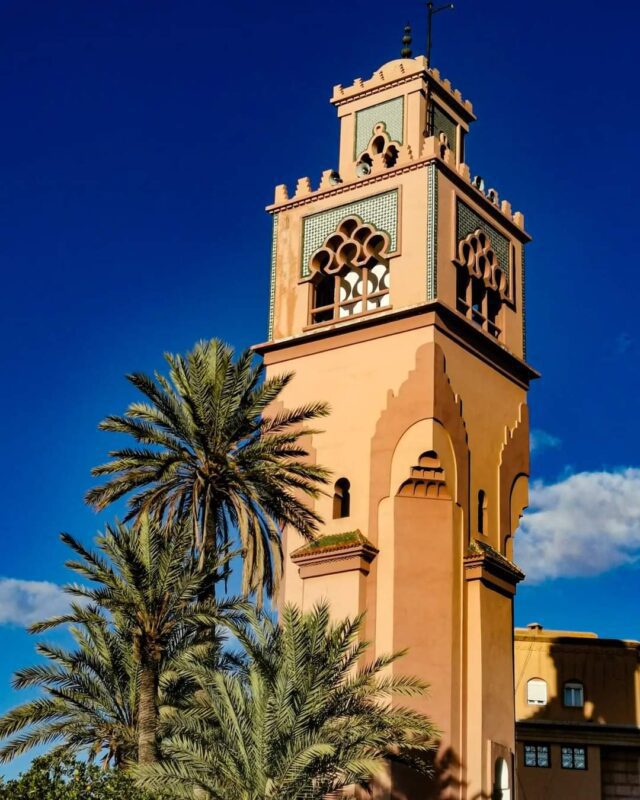 Mosque minaret with carved arches and green tilework rising above palm trees against a clear blue sky
