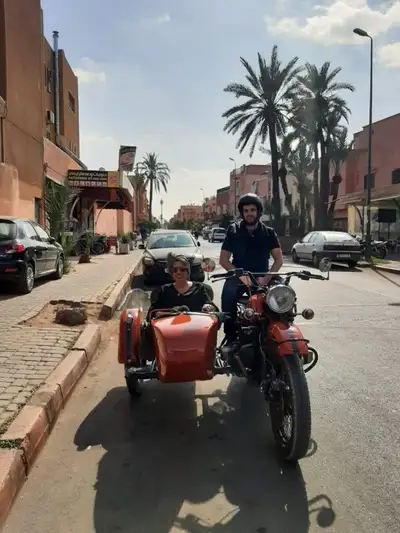 Couple riding an orange vintage motorcycle with sidecar past palm-lined streets in Marrakech