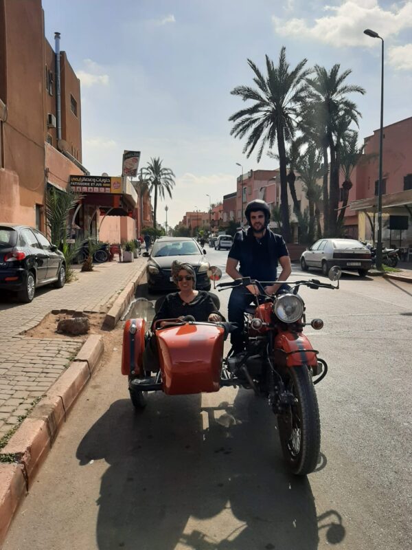 Couple riding an orange vintage motorcycle with sidecar past palm-lined streets in Marrakech