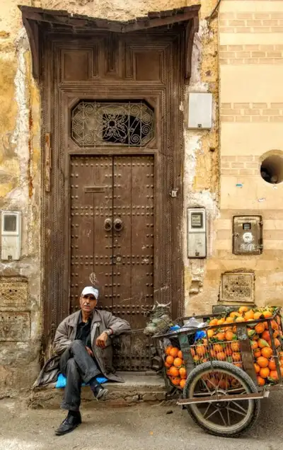 Man sitting beside a wooden cart heaped with oranges in front of a studded medina door