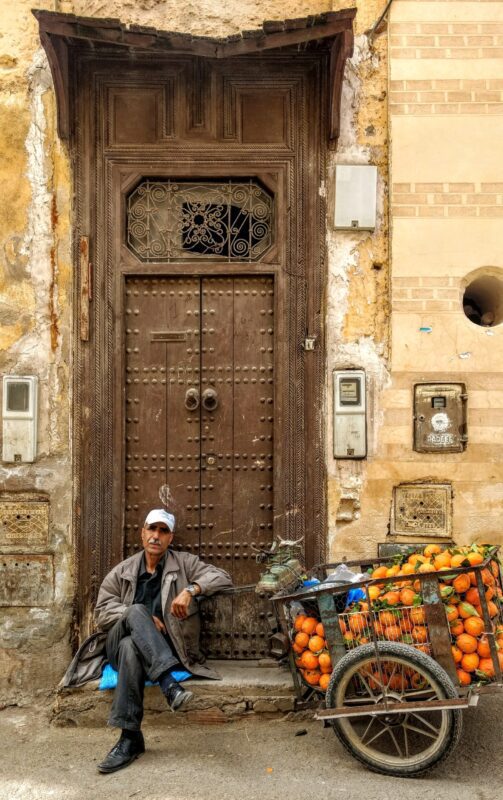 Man sitting beside a wooden cart heaped with oranges in front of a studded medina door