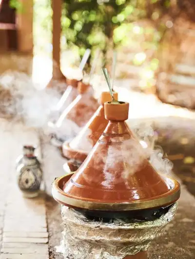 Row of steaming terracotta tagines cooking outdoors on foil-wrapped charcoal burners